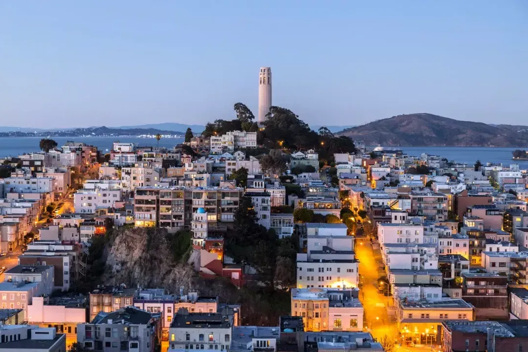 Coit Tower de San Francisco al anochecer, con calles iluminadas frente a ella y la Bahía de San Francisco detrás.