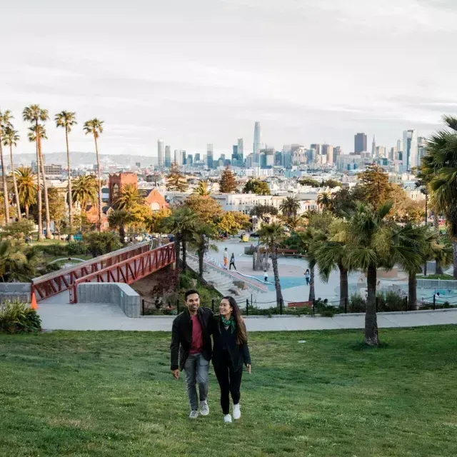 Un couple marche vers la caméra avec Dolores Park et la skyline de San Francisco derrière eux.